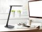 Preview: Moderner Arbeitsplatz mit schwarzer Schreibtischlampe, Computerbildschirm, Kaffeetasse, Brille und Smartphone auf weißem Tisch vor Fenster mit Pflanzen.