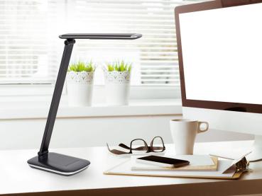 Preview: Moderner Arbeitsplatz mit schwarzer Schreibtischlampe, Computerbildschirm, Kaffeetasse, Brille und Smartphone auf weißem Tisch vor Fenster mit Pflanzen.